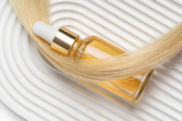 A luxurious golden hair oil in a clear dropper bottle rests on blonde hair against a wavy white background, symbolizing upscale hair care