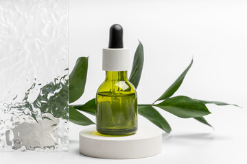 A clear glass dropper bottle filled with green botanical oil, presented on a simple white pedestal with green leaves, against a textured backdrop