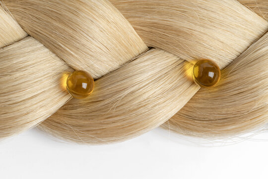 A close-up of a beautifully woven blonde hair pattern with two translucent amber-colored oil capsules placed strategically, showcasing a luxurious hair treatment