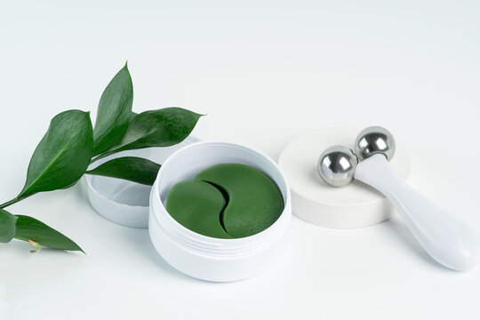 A wellness setup featuring green under-eye patches in a jar and a white facial roller with metallic balls, accented by fresh green leaves