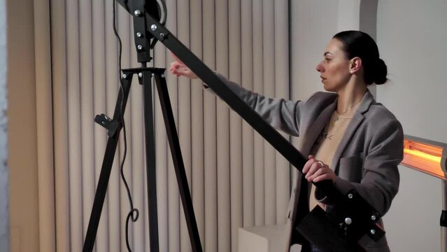 A young woman photographer in a gray suit adjusts professional lighting equipment for an indoor photo shoot. She is seen adjusting the height of a tripod and positioning a halogen lamp. 