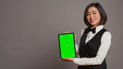 Woman concierge presenting greenscreen display on tablet, showing chromakey template on screen. Receptionist with bow and uniform holding device with blank copyspace layout. Camera B.