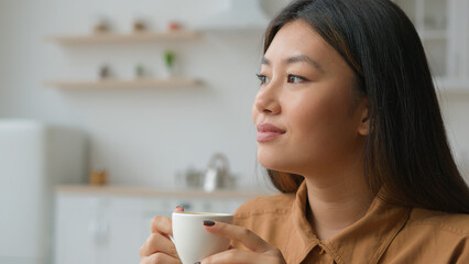 Peaceful relaxed Asian chinese japanese woman sit in kitchen hold cup of tea dreaming think feel...