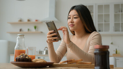 Asian woman in kitchen use smartphone mobile phone application surfing internet. Multiracial korean...