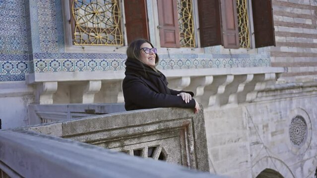 A smiling woman in a scarf leaning on the balcony of topkapi palace in istanbul, showcasing traditional architecture.