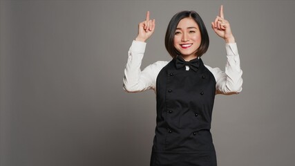 Asian woman pointing at something upwards in studio, indicating direction above her head. Waitress...