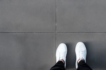 A person's feet clad in clean white sneakers, positioned on a textured grey pavement. White Sneakers Standing on Grey Pavement - Powered by Adobe