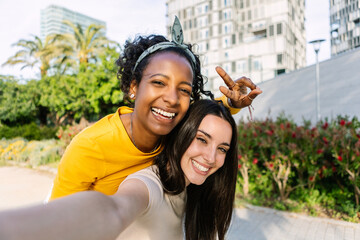 Two young beautiful women taking selfie portrait together in summer at city. Diverse girls enjoying...