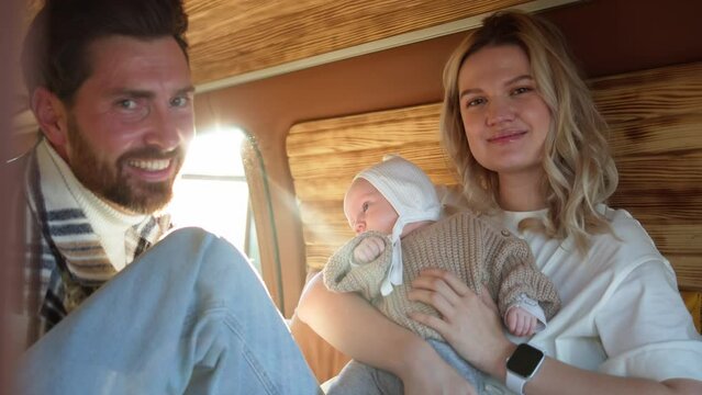 A smiling man and woman adoring their serene baby wrapped in a knitted sweater inside a cozy camper van.