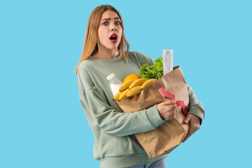 Shocked young woman with arrow and bag of food on blue background. Price rise concept