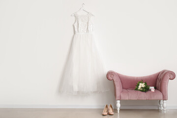 Wedding dress hanging on white wall and bridal bouquet on pink velvet armchair in room