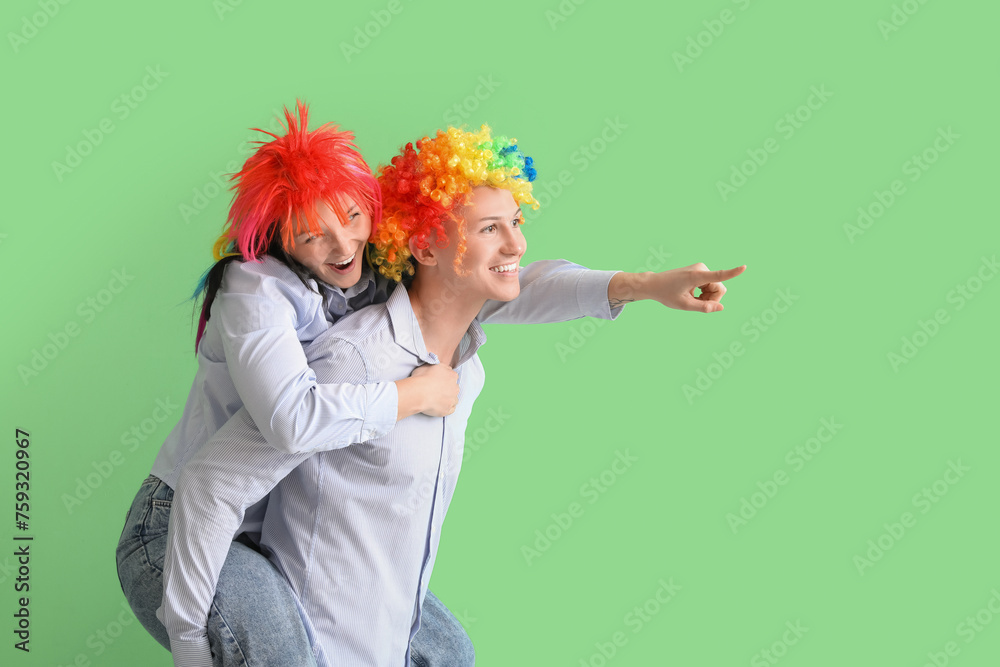 Poster Business colleagues in funny wigs pointing at something on green background. April Fools' Day celebration