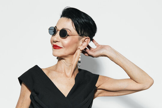 Stylish older woman wearing sunglasses and black dress posing for camera in front of white wall
