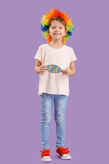 Cute little boy with colorful clown wig and paper fish on lilac background. April Fool's Day celebration