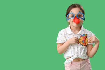 Cute little girl with funny disguise and paper fish on green background. April Fool's Day celebration - obrazy, fototapety, plakaty