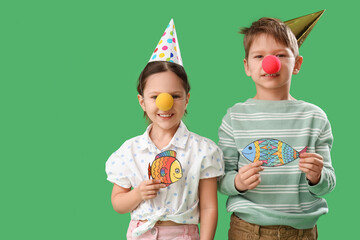 Little brother and sister with clown noses, party hats and paper fishes on green background. April Fool's Day celebration - obrazy, fototapety, plakaty