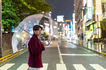 Portrait of Young Asian man holding umbrella walking city street crosswalk with night lights in Tokyo city, Japan in raining night. Loneliness guy walking in the city at night.