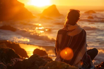  A person sitting on rocks at the beach, watching the sunset over the ocean in California, with warm colors of the sunrise © artfisss