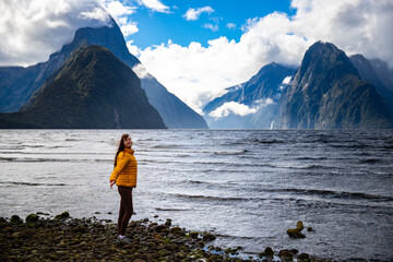 Happy Girl in yellow jacket dances and admires famous Milford Sound fiord and mighty mountains densely covered with vegetation in Fiordland National Park, Southland, New Zealand