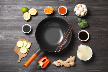 Empty iron wok and chopsticks surrounded by ingredients on dark grey wooden table, flat lay