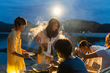 Group of Young Asian man and woman friends enjoy and fun celebration meeting dinner party eating barbecue grill and drinking beer together on lake house balcony on summer holiday vacation at night.