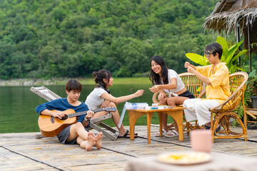 Young Asian man and woman enjoy outdoor lifestyle travel nature forest mountain on summer holiday vacation. Happy generation z people friends playing guitar and doing hobbies at lake house balcony.