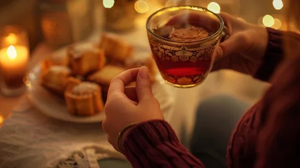 Tafelkleed A close-up of a woman's hands holding a cup of tea, with a plate of Ramadan sweets in the background, © Daunhijauxx