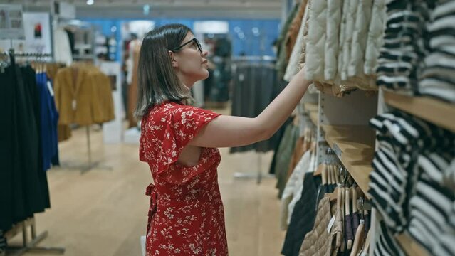 Fashion passion! beautiful hispanic woman with glasses choosing chic clothes in a retail store. portrait of a young, brunette shopper shopping at a boutique in the mall.