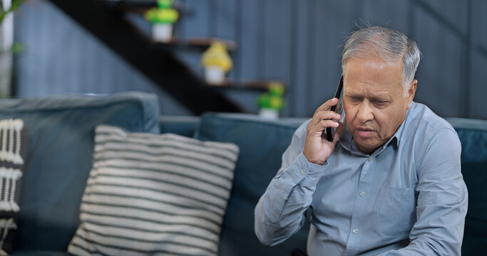 Indian Asian Hindu Upset tensed angry stress old elder man sitting sofa couch talk share issue shock news phone call use mobile alone aged older sad gen x male guy feel bad indoor room home house