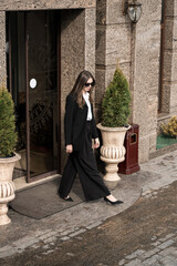 A stylish woman in a black suit and white shirt with glasses poses for the camera against the...