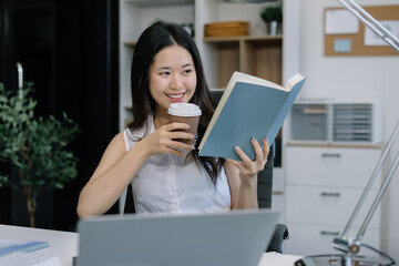Asian businesswoman reading a book sits in a office and enjoys a coffee.