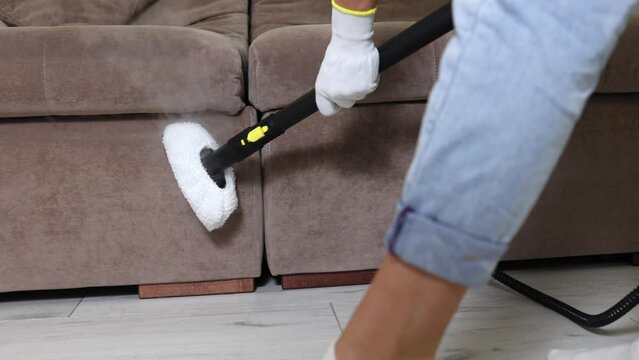 The hand of employee of a cleaning company in protective glove cleans side soft surfaces of a brown sofa in an apartment with a steam mop. Wet Professional Cleaning of Home Upholstered Furniture