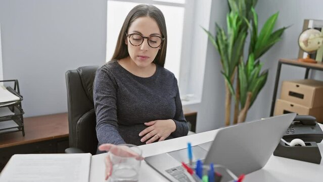 A pregnant hispanic woman wearing glasses drinks water while working at her office desk.