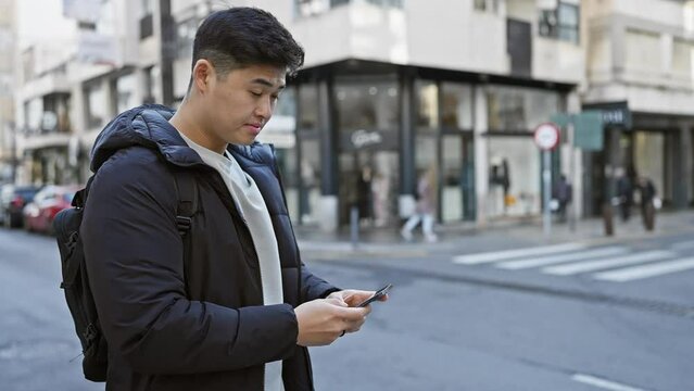 Young asian man using smartphone on busy city street with blurred pedestrians and urban backdrop.