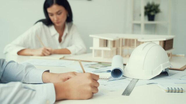 Professional architect engineer drawing blueprint and working together with young beautiful caucasian coworker on meeting table with safety helmet, house model, blueprint scatter around. Immaculate.