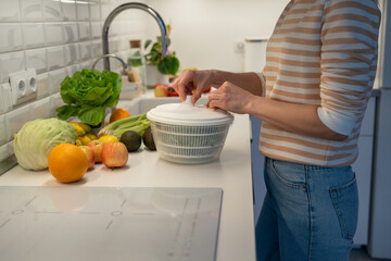 Slim vegan woman preparing useful dinner of fresh fruit and vegetables. Concentrated female drying greenery in plastic hand food centrifuge. Tasty ripe oranges, apples. cabbage, avocado lying on table