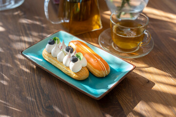 Two handmade eclairs are displayed on a ceramic plate standing on a wooden table. eclair with white cream and blueberries, the second in orange glaze. Glass tea set in the background.