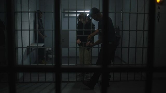 Warden takes off handcuffs from young prisoner standing behind metal bars. Multi ethnic teenagers serve imprisonment term in correctional facility or detention center. Guilty inmates in prison cells.