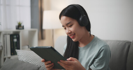 Selective focus, Young asian woman wear wireless headphones sitting on sofa using digital tablet for online shopping cashless in living room at home - 759606150