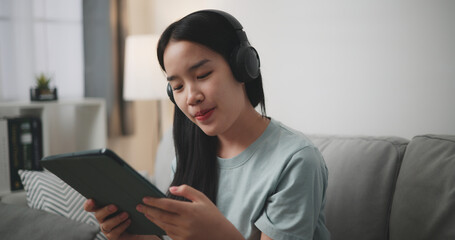 Selective focus, Young asian woman wear wireless headphones sitting on sofa using digital tablet...