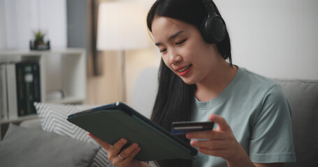 Selective focus, Happy young woman wear wireless headphones sitting on sofa using digital tablet and credit card for online shopping cashless in living room at home - 759606155