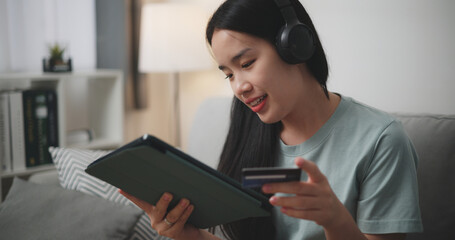 Selective focus, Happy young woman wear wireless headphones sitting on sofa using digital tablet and credit card for online shopping cashless in living room at home - 759606173
