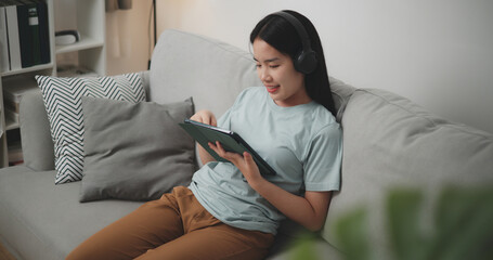 High angle view, Young asian woman wear wireless headphones sitting on sofa using digital tablet for online shopping cashless in living room at home - 759606316