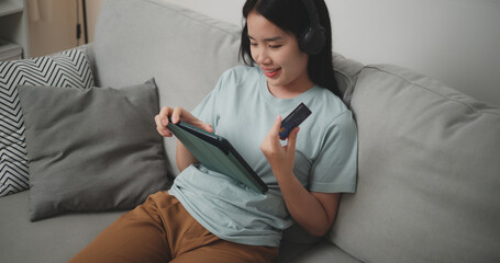 High angle view, Happy young woman wear wireless headphones sitting on sofa using digital tablet and credit card for online shopping cashless in living room at home - 759606353