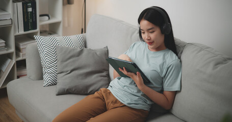 High angle view, Young asian woman wear wireless headphones sitting on sofa using digital tablet for online shopping cashless in living room at home - 759606364
