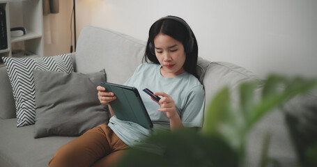 High angle view, Happy young woman wear wireless headphones sitting on sofa using digital tablet and credit card for online shopping cashless in living room at home - 759606387