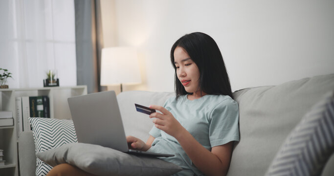 Selective focus, Young asian woman sitting on sofa holding credit card making online payment for purchase in web store using laptop.