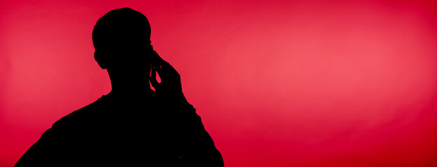 dark silhouette of a male student talking with a smartphone on a pink background, close-up with...
