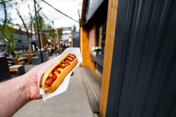  hand holding a hotdog with generous ketchup and mustard toppings, set against an urban backdrop. Outdoor seating arrangements and string lights create an inviting atmosphere