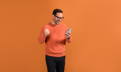Overjoyed young male winner shaking fist and reading good news over smart phone on orange background